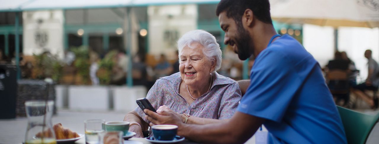 Senor woman having coffee with a male nurse while looking at  something on a mobile phone.