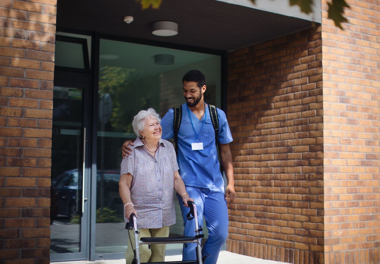 Senior woman with her walker exits a building accompanied by a male nurse.