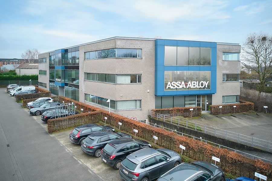 ASSA ABLOY Entrance Systems gebouw in Melle met groot ASSA ABLOY-logo op het raam van de tweede verdieping, omringd door groen en een parkeerterrein met auto's.