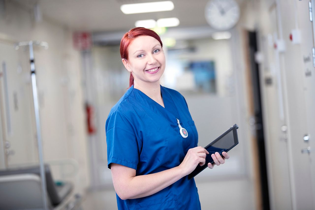 Nurse standing in a hospital corridor holding a tablet.