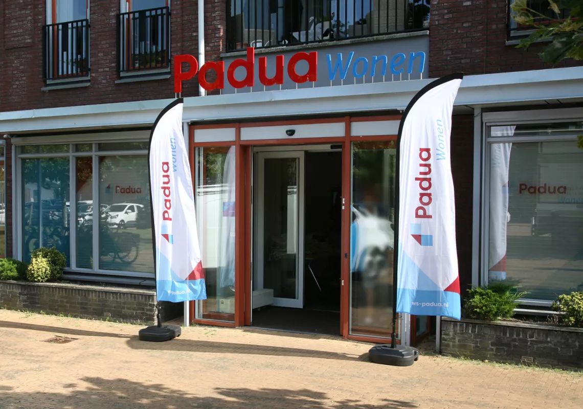 Ingang van een gebouw met twee beachflags ervoor en een bord erboven met de tekst Padua Wonen.