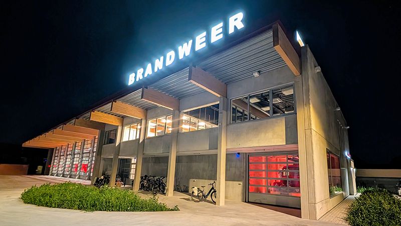 Photo prise le soir depuis le parking de la caserne des pompiers de Zele, avec tous les éclairages allumés et les camions de pompiers visibles derrière les portes sectionnelles vitrées.