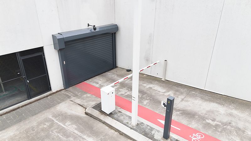 Porte de parking gris foncé dans le parking souterrain de Fluvius à Melle, avec bande cyclable rouge, barrière et lecteur de badge.