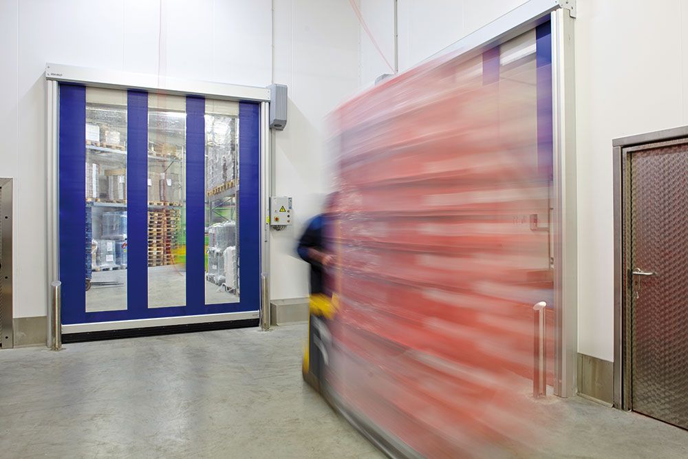 Une personne conduit un chariot élévateur à fourche dans une zone de stockage remplie de marchandises, avec en arrière-plan un portail robuste de voie rapide