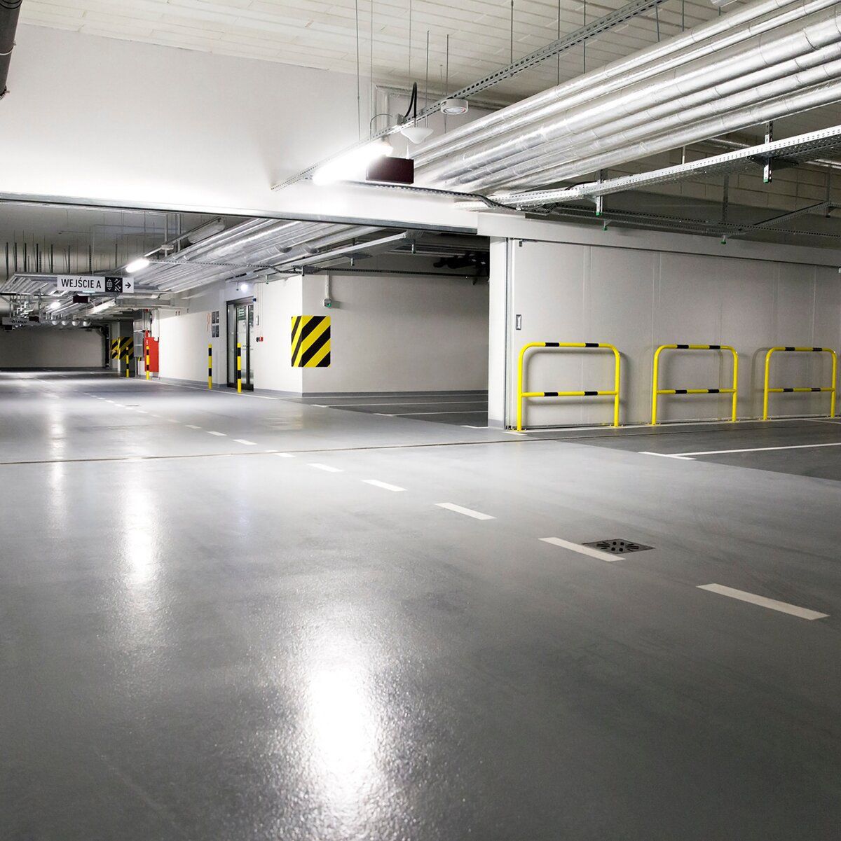 En underjordisk parkeringskælder med en brandklassificeret porte, glat reflekterende gulv, advarselsskilte, gule afspærringer og rørledninger i loftet.