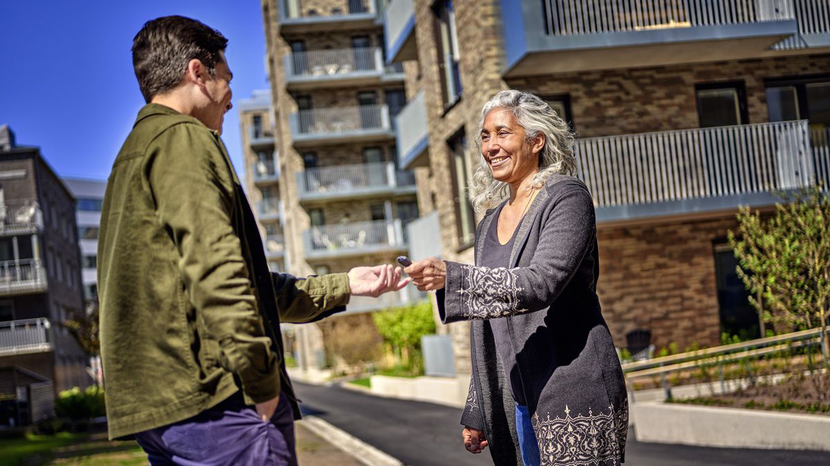 Kvinna tar emot en ASSA ABLOY PULSE-nyckel från en man ute på gården i ett flerbostadshusområde.