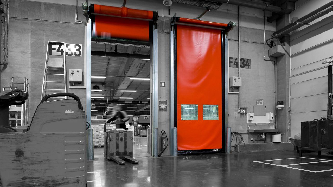 Two red slim high speed doors, and a black and white warehouse setting with a worker driving through one of the doors. 
