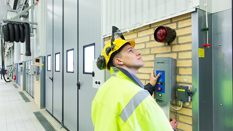 Technieker in veiligheidsuitrusting (helm, bril, fluohesje) inspecteert een sectionale poort in een industrieel gebouw en bedient de controlekast om een storing te analyseren.