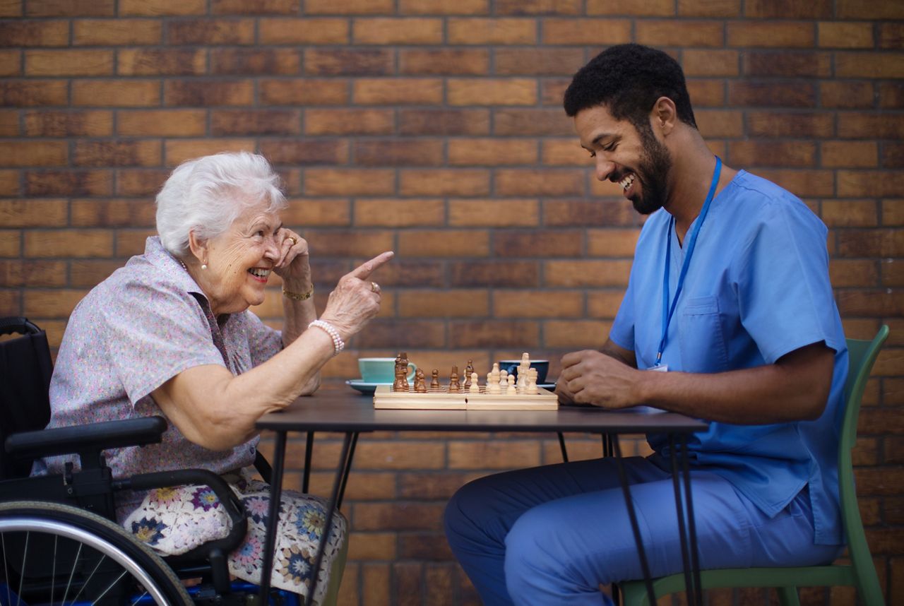 Äldre dam sitter och spelar schack ute ihop med manlig sköterska.