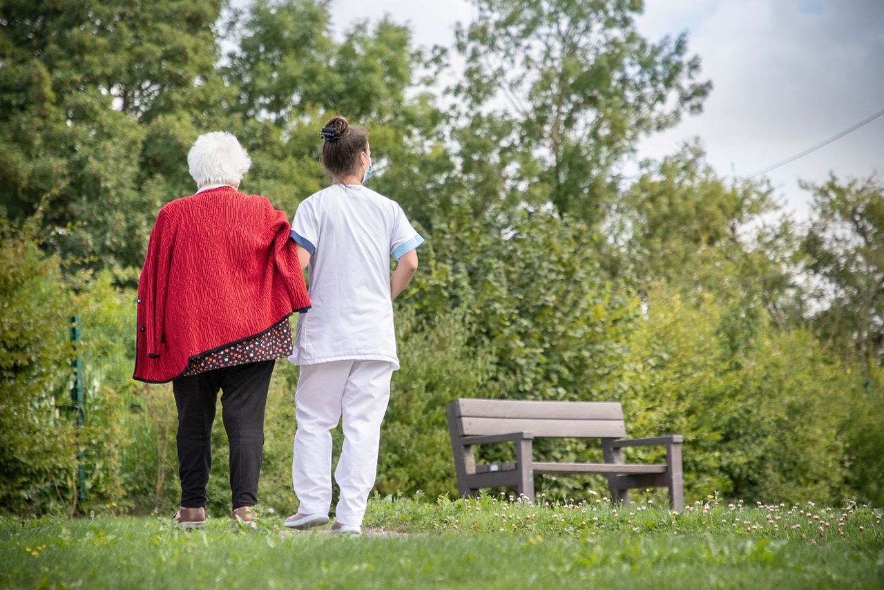 Sygeplejerske, der går i en park med ældre kvinde i rød jakke.