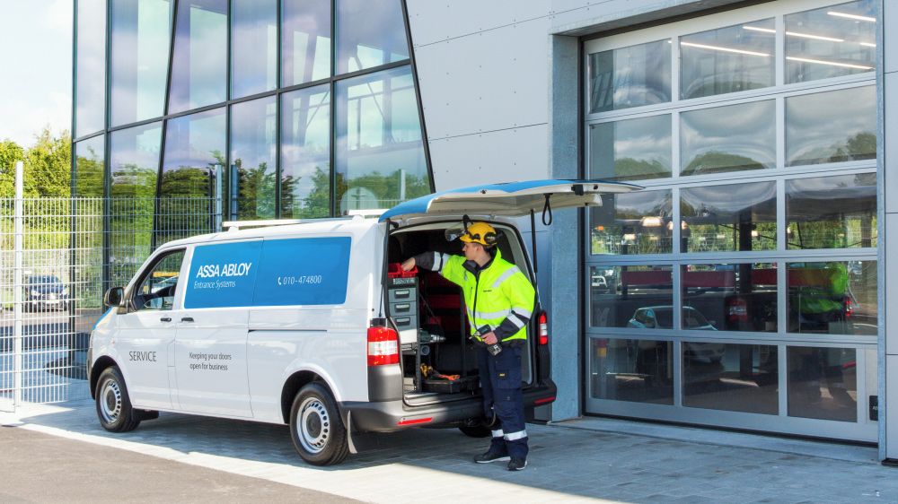 Un homme en tenue de travail avec gilet fluorescent et casque retire un équipement de travail d'une camionnette ASSA ABLOY pour une intervention technique.