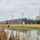 Un long bâtiment moderne avec des façades en bois et sombres, situé le long d'un front de mer avec des roseaux et de jeunes arbres.