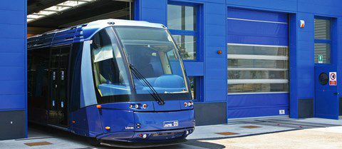 Modrá tramvaj vyjíždějící z depa, vpravo jsou modrá průmyslová vrata