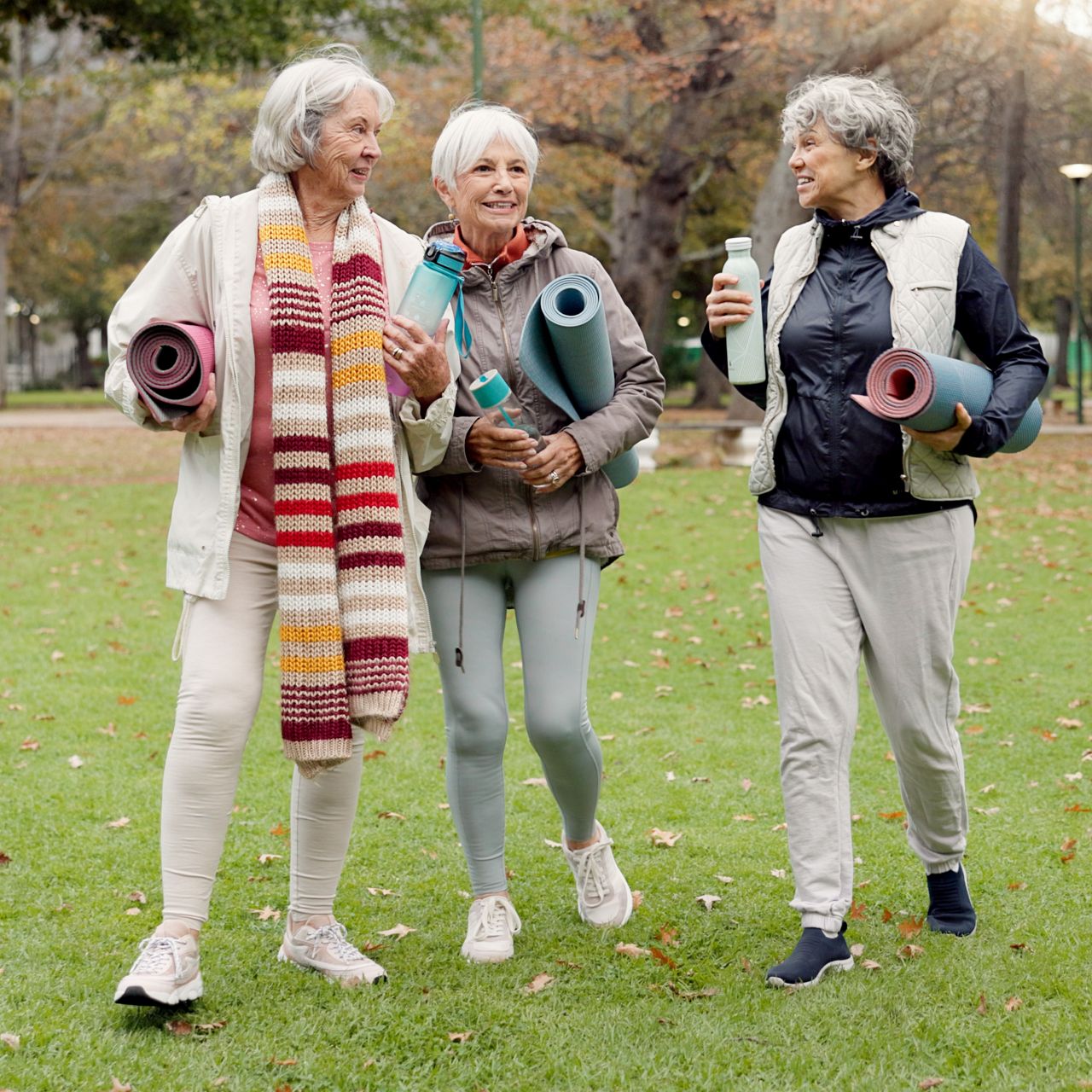 Three women of senior age carrying yoga mats in a park.