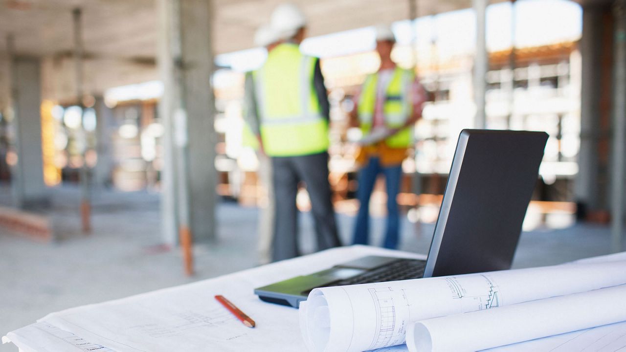 View of a construction site, in the foreground is a table with drawings and an opened laptop, in the background is three people in yellow vests and safety helmets.