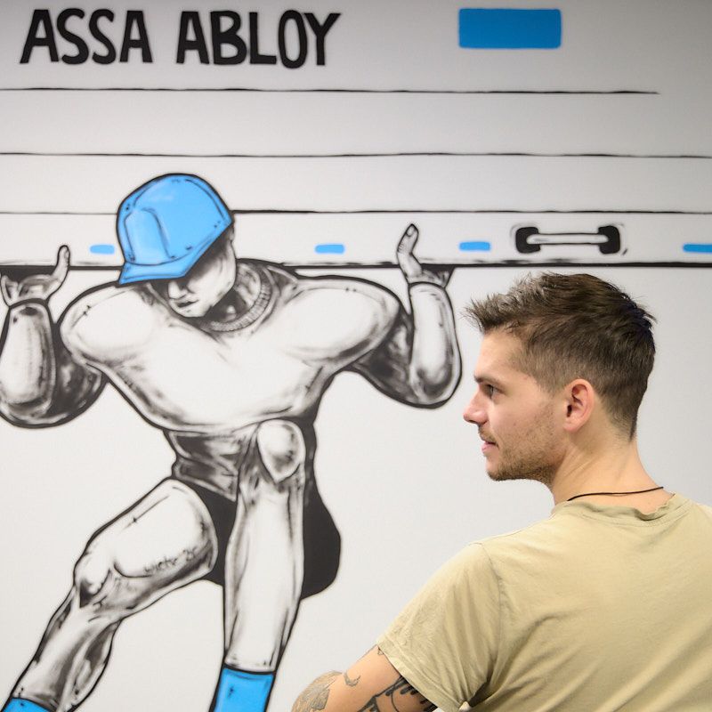 Streetartkunstenaar Wietse zit ontspannen in een zetel en kijkt naar zijn net afgewerkte muurschildering in het ASSA ABLOY-kantoor, waarop een technieker te zien is die een poort optilt.